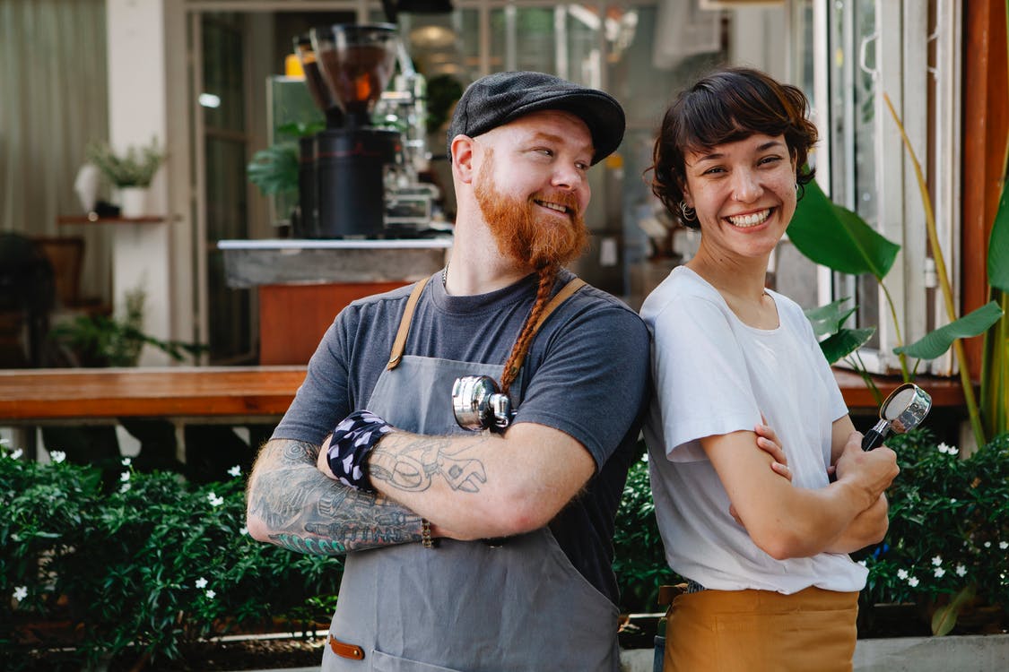Couple of smiling barista coworkers in casual outfit and aprons standing with portafilter near cafeteria in street near plants in daylight