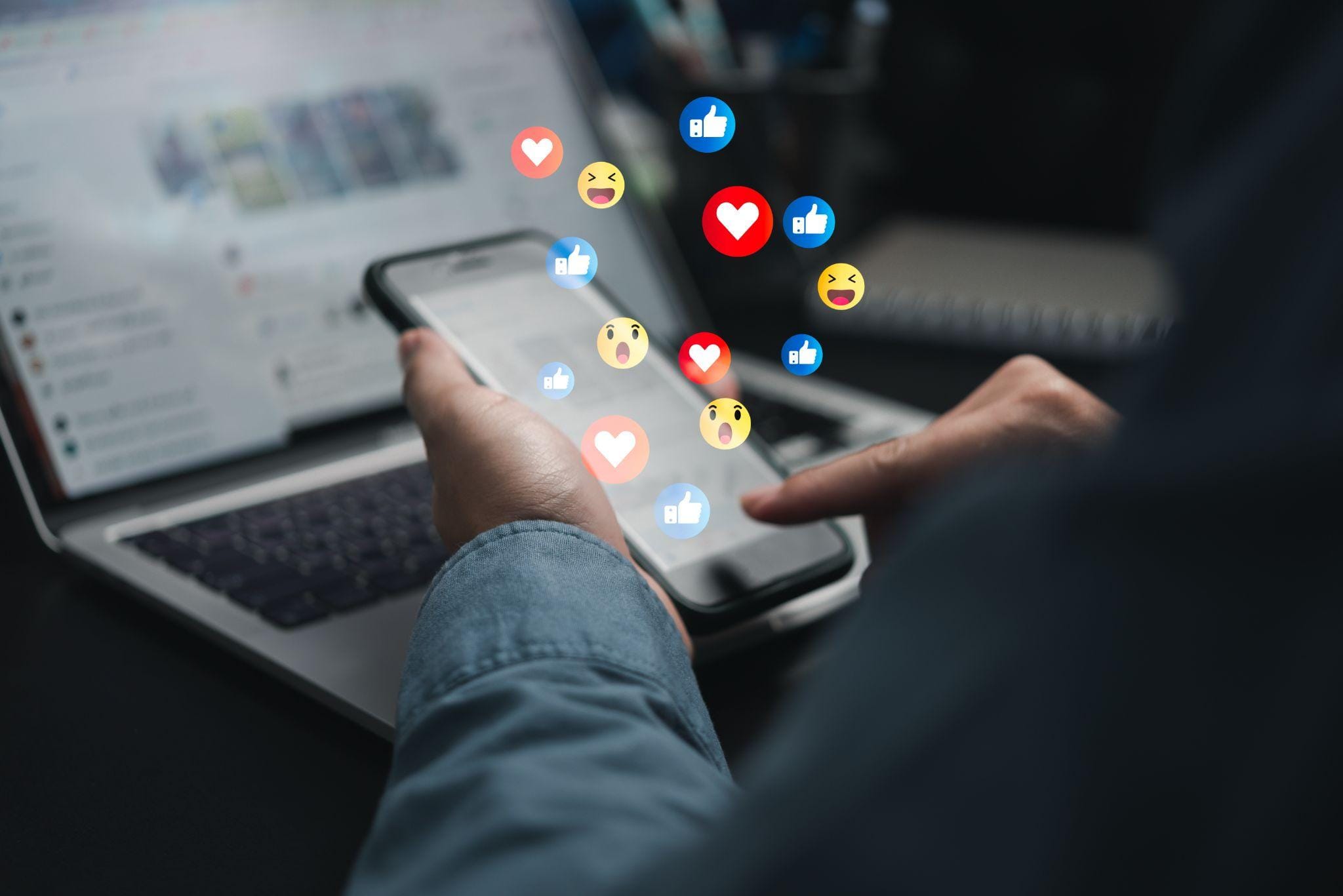 Person using a smartphone with floating social media reaction icons, including likes and hearts, while working on a laptop, representing digital marketing and social media engagement.